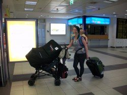 Ich mit Tochter auf dem Rücken, Hüfttasche mit Papieren, Wickeltasche als Handgepäck am Buggy, auf dem Buggy den Autositz im Airshell und eine Rolltasche